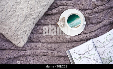 Weiße Tasse Kaffee mit Bild Strand mit Meer Wellen und Waffel Cookie in Form Tanne, weiße Seiten Buch mit Karte reisen auf Kissen und warme Wolldecke Grau karierte. Winter herbst Konzept Komfort und Träume. Stockfoto