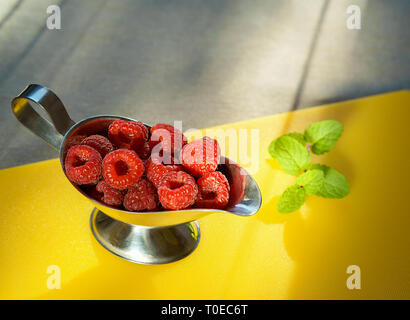 Süße Himbeeren in Silver Bowl und Minze auf gelbe Küche board Reif im Licht der Sonne. Nahaufnahme, Ansicht von oben, gesundes Frühstück Zutat Stockfoto