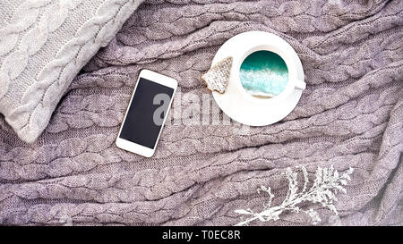 Weiße Tasse Kaffee mit türkisblauem Meer Wellen und Waffel Cookie in Form Tanne und weiß Telefon Blume auf Kissen und warme Wolldecke Grau karierte. Gestrickte textile mit braid Muster. Winter herbst Konzept Komfort Stockfoto