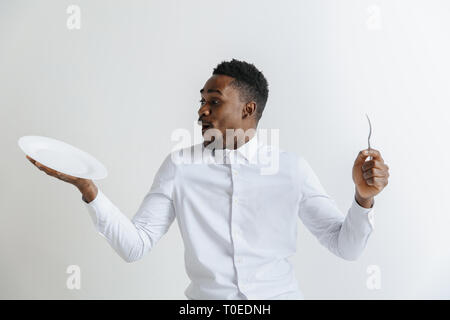 Junge überrascht attraktive african american guy Holding leere Teller und Gabel auf grauem Hintergrund. Kopieren Sie Raum und Mock up. Leere Vorlage Hintergrund. Stockfoto