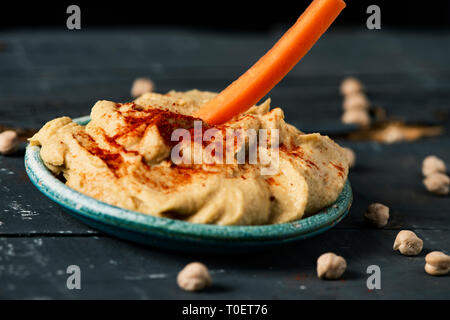 Nahaufnahme von einem Streifen von Karotte in einem hausgemachten Hummus gewürzt mit Paprika in einer grünen Keramik Teller getaucht wird, auf einem dunklen Grau rustikal Tab Stockfoto