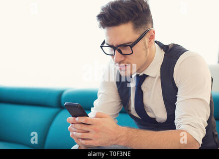 Junge Unternehmer in Gläsern sitzen auf der Couch mit Smartphone suchen aufgeregt und verwirrt Stockfoto