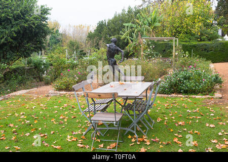 Herbst oder Fallen Rosengarten mit Blätter auf dem Rasen und über Holz- leeren Tisch und Stühlen und im Hintergrund eine schöne Statue oder Skulptur verstreut Stockfoto