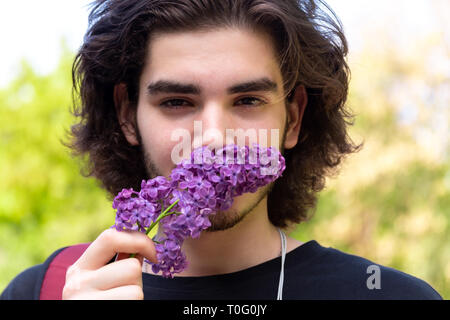 Junge brunette Kerl Duftende frische Blumen auf lila Zweig und Kamera auf unscharfen Hintergrund Stockfoto