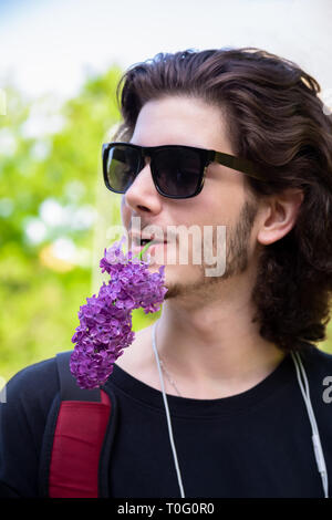 Junge brunette Mann mit langen Locken und Sonnenbrille hält frische Blumen in seinen Mund auf einem lila Zweig und schaut weg auf einem verschwommenen Hintergrund. Stockfoto