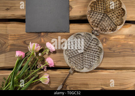 Traditionelle alte Waffeleisen auf rustikale Holzplanken mit Schiefer und Blumen angeordnet. Von oben, flach Schuß. Stockfoto