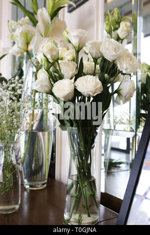 Ein Glas Vasen aus weißem lisianthus, Gypsophila und Lilien auf dem Sideboard Schrank in der Nähe von einem Fenster im Wohnzimmer setzen Stockfoto