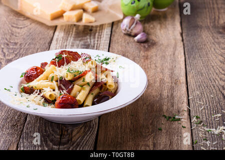 Rigatoni Nudeln mit Salami, gebratene Tomaten und Oliven, serviert köstliche italienische Gerichte mit geriebenem Parmesan auf rustikalen Holztisch Stockfoto