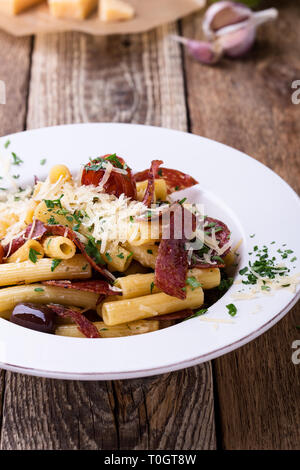 Rigatoni Nudeln mit Salami, gebratene Tomaten und Oliven, serviert köstliche italienische Gerichte mit geriebenem Parmesan auf rustikalen Holztisch Stockfoto