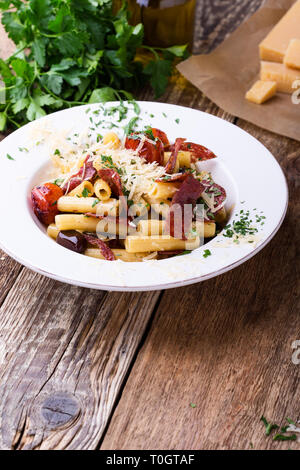 Rigatoni Nudeln mit Salami, gebratene Tomaten und Oliven, serviert köstliche italienische Gerichte mit geriebenem Parmesan auf rustikalen Holztisch Stockfoto