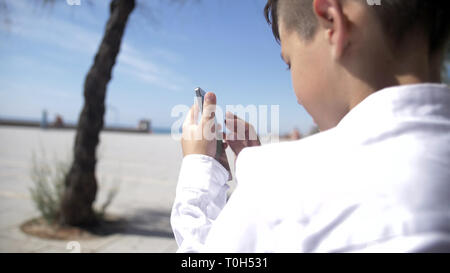 Junge sitzt auf einer Bank vor dem Hintergrund der Strand und verwendet das Telefon, haben Spaß Stockfoto