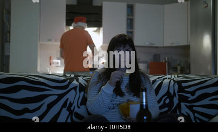 Frau vor dem Fernseher, trinkt Bier mit Chips, ein mann Essen bereitet Stockfoto