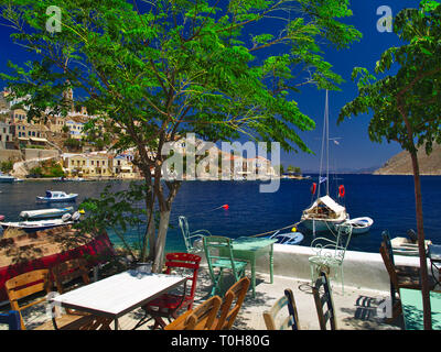 Ein Sommer Schuß von der Insel Symi in Griechenland. Blick von einem Café mit Tischen, Sesseln Boote kristallklaren Meer und den bunten Häusern. Stockfoto