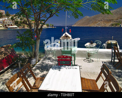 Ein Sommer Schuß von der Insel Symi in Griechenland. Blick von einem Café mit Tischen, Sesseln Boote kristallklaren Meer und den bunten Häusern. Stockfoto