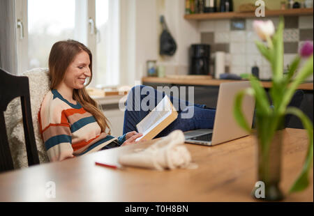 Schöne blonde Frau an einem Tisch sitzen, ein Buch zu lesen, lächelnd, Laptop und häkeln Utensilien auf dem Tisch Stockfoto