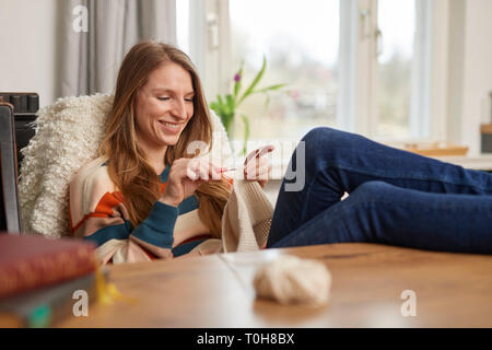 Schöne blonde Frau sitzt an einem Tisch häkelt mit weißem Garn an der Nadel lächelnd Stockfoto