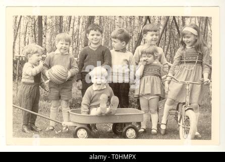 Original Postkarte aus den 1960er Jahren mit einer Gruppe glücklicher junger britischer Kinder, Jungen und Mädchen sowie Kleinkindern im Alter von 2 bis 4 Jahren, die im Freien spielen, mit Fahrrad und Zugwagen, (Generation X Gen X oder Baby Boomers) Großbritannien Stockfoto