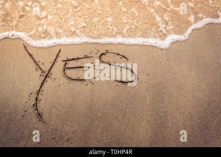 Das Wort "ja" handschriftlich auf dem Sand von einem Strand bei Sonnenuntergang mit Wellen auf das Ufer Stockfoto