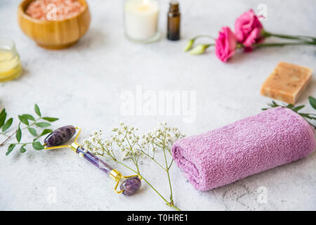 Spa Noch leben Einstellung mit Amethyst Gesicht Roller, Blumen, Handtuch und natürlichen Home Spa Produkte auf konkreten Hintergrund, selektiver Fokus, Spa & wellne Stockfoto