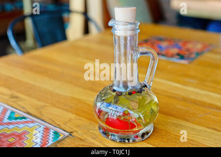 Ein Glas Flasche frisches Olivenöl mit ganzen Pfeffer in Hühneraugen und einige Kräuter auf einem Tisch. Niemand um Stockfoto