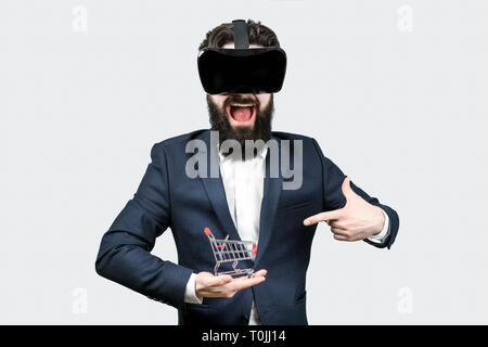 Der bärtige Mann überrascht in der virtuellen Realität Brille weg zeigend auf einen Warenkorb, virtuelle Shopping Konzept Stockfoto