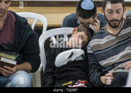 Elkana, Israel. 20 Mär, 2019. Ein Junge mit einem phanton der Oper Gesichtsmaske auf seiner Brust und einer Spielzeugpistole auf seine Knie, schläft während der Lesung des Blättern von Esther in der Synagoge in der israelischen Ortschaft Elkana während der jüdische Feiertag Purim. In diesem Urlaub Juden traditionell verkleiden sich, lesen Sie das Buch von Esther, eine Geschichte über ein anti-jüdischen fehlgeschlagen Grundstück im alten Persien, und, wann immer ein Schläger der Name Haman, der main Antagonist, erwähnt wird. Credit: Yagil Henkin/Alamy leben Nachrichten Stockfoto