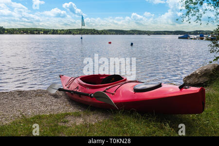 Rotes Wasser reisen Kajak liegen auf dem Strand von Wörthsee. Im Hintergrund die See mit Flagge, Boote und Pier in Wörthsee, Bayern, Deutschland, Europa Stockfoto