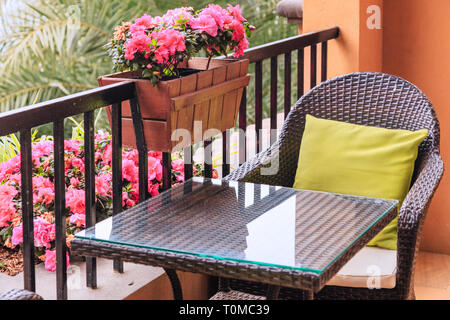 Ratten Tisch und Sessel mit Kissen auf dem Balkon, durch vergossene Blüten rosa Azalee auf der Balustrade mit grünen Palmen im Hintergrund. Stockfoto