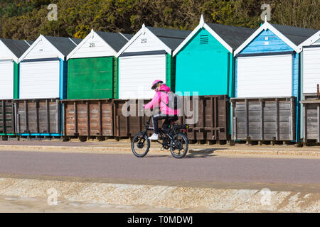 Junge Frau, Radfahren, Reiten ein eco Servolenkung elektrische Fahrrad entlang der Promenade Vergangenheit Holzhütten am Bournemouth, Dorset UK im März Stockfoto