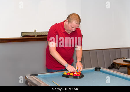 Ein Mann mittleren Alters in einem roten T-Shirt stellt die Bälle für eine Partie Billard auf dem blauen Baize-Tisch auf. Stockfoto