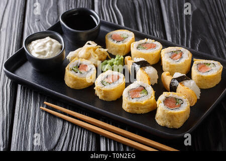 Tamagoyaki Omelett set Sushi Rollen mit Reis, geräuchertem Lachs und Avocado close-up mit Soßen, Wasabi und Ingwer auf einer Platte auf einem Schwarzen Tisch. Horizont Stockfoto