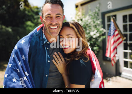 Lächeln, Mann und Frau, die in ihrem Hinterhof mit der amerikanischen Flagge im Hintergrund. Glückliches Paar stehen zusammen mit der amerikanischen Flagge auf Th Stockfoto