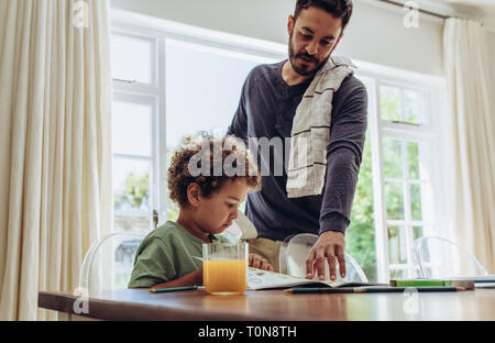 Junge sitzt am Tisch ein Buch lesen mit einem Glas Saft auf dem Tisch. Der Mensch auf der Suche nach seinem Kind und täglichen Haushalt arbeitet. Stockfoto