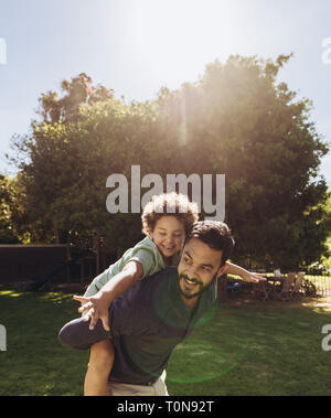 Man Walking im Park seinen Sohn auf dem Rücken auf einem sonnigen Tag. Glückliches Kind genießen auf dem Rücken seines Vaters mit offenen Armen. Stockfoto