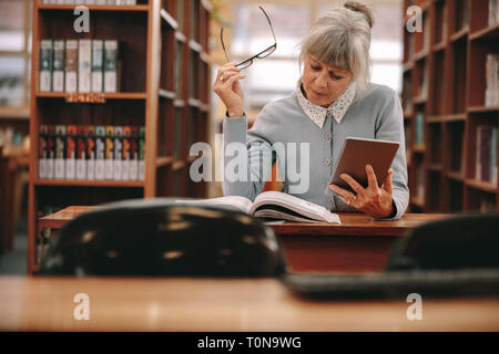 Ältere Frau mit einem Tablet-PC als Referenz mit einem Buch in der Bibliothek. Frau sitzt in einer Bibliothek ein Buch lesen. Stockfoto