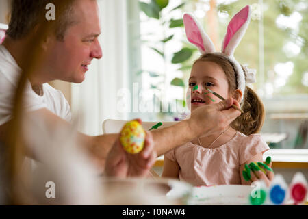 Ein Vater und eine Tochter Ostern feiern, malen Eier mit der Bürste. Glückliche Familie zu lächeln und zu lachen, Zeichnung im Gesicht. Süße kleine Mädchen im bunny Ohren Stockfoto