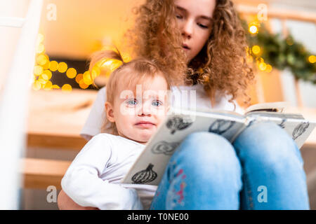 Ältere Schwester lesen Buch vorlesen zu ihrer jüngeren Schwester an der Treppe. Girlande Lichter auf Hintergrund. Familie Liebe und Unterstützung. Stockfoto