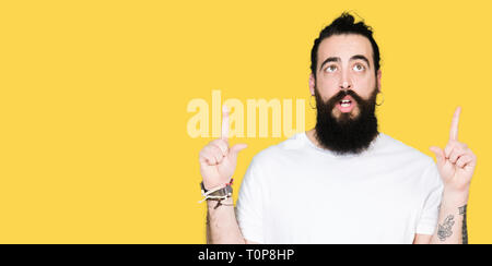 Junge hipster Mann mit langen Haaren und Bart legere weißes T-Shirt erstaunt und mit der Suche und zeigen mit den Fingern überrascht und erhobenen Armen. Stockfoto