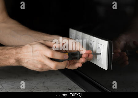 Nahaufnahme der Hand ein Elektriker mit einem Schraubendreher Auseinandernehmen eines weißen Steckdose auf eine schwarze Glaswand. Stockfoto