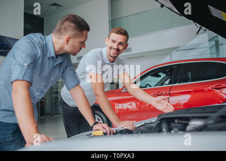 In weissen Sirt steht Verkäufer bei eröffnet Körper von Auto und Punkt. Er schaut auf das Kerl und lächelt. Mann im grauen Shirt lehnt sich an Auto und schaut auf den Verkäufer. Er Stockfoto