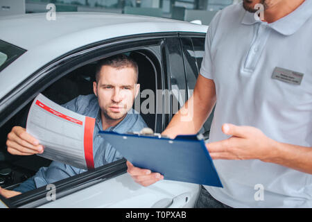 Verkäufer steht an weißes Auto und hält Tablett in den Händen. Er Punkt auf. Ein anderer Mann sitzt im Auto und hält Versicherung Papier. Er ist ernst und konzentrieren Stockfoto