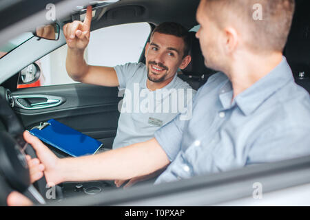 Junge und bärtige Berater sitzen im Auto. Er schaut auf Kunden- und nach oben zeigt. Junger Mann lächelt. Käufer sitzt auf der vorderen Sitzen und Verkäufer. Er halten Stockfoto