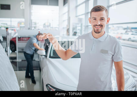 Bild von Auto deeler steht und schaut auf Kamera. Er lächelt und hält Auto-Taste. Potenzielle Käufer steht und lehnt sich auf weißen Auto. Er sieht in der i Stockfoto