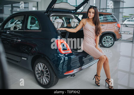 Wunderschöne und selbstbewusste Modell sitzt am Rande der Amtsleitung. Sie posiert und schaut auf Kamera. Junge Frau berührt Auto mit der Hand. Sie trägt Kleid und Schuhe auf Hi Stockfoto