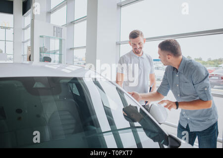 Junger Mann stand vor der weißen Auto und es berührt. Alo blickt er auf Verkäufer und mit ihm zu sprechen. Junger bärtiger Mann schau es Dir an und lächelt. Sie sind im Auto. Stockfoto