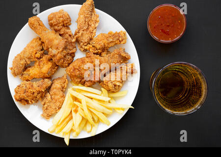 Lecker Fastfood: Fried Chicken Drumsticks, würzige Flügel, Pommes frites, Chicken Fingers, Glas kaltes Bier, Soße auf weiße Platte über schwarze Oberfläche, oben Stockfoto