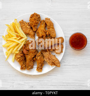 Lecker Fastfood: Gebratene Hähnchenschenkel, würzige Flügel, Pommes frites, Hähnchenstreifen und sauer - süsse Soße auf weiße Platte über weiß Holz- Hintergrund, oben vi. Stockfoto