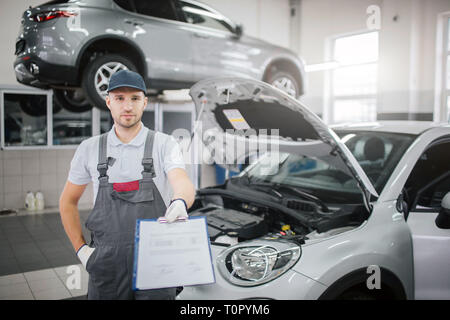 Schön und selbstbewussten jungen Mensch steht im Auto und schaut auf Kamera. Er hält Plastik Ordner mit Dokumenten auf. Eine Hand in der Tasche. Körper des Auto ist op Stockfoto