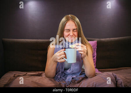 Schöne junge Frau schrumpft von Zitrone. Sie hält Stück davon in der einen Hand und heiße Tasse Tee in eine andere. Frau leidet. Stockfoto