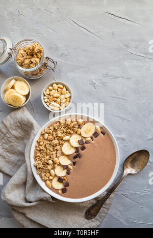 Schokolade smoothie Schüssel mit Bananen, Müsli und Erdnüsse auf Hellgrau konkreten Hintergrund. Gesundes vegetarisches Frühstück. Ansicht von oben. Stockfoto